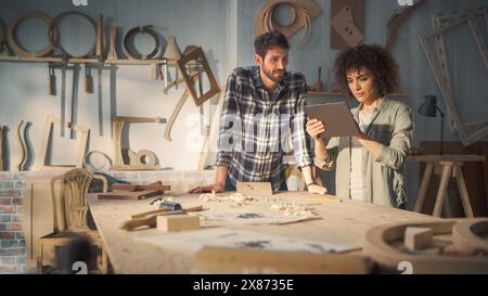 Zwei junge Kleinunternehmer, die einen Tablet-Computer verwenden und über das Design eines neuen Holzmöbelprojekts sprechen. Zimmermann und eine junge weibliche Auszubildende, die im Loft Studio arbeitet. Stockfoto