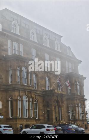 Ein historisches Hotelgebäude in nebeliger Atmosphäre mit mehreren Bogenfenstern, zwei Flaggen und davor geparkten Autos. Stockfoto