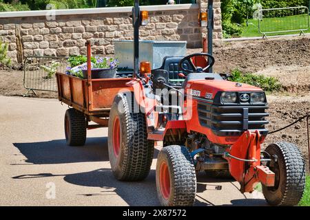 Ein roter Traktor mit einem Anhänger mit Topfpflanzen, der auf einem gepflasterten Weg in einem Gartenbereich abgestellt ist. Stockfoto