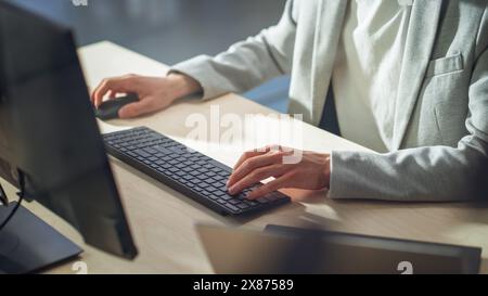 Junger männlicher Mitarbeiter in grauer Freizeitjacke, der tagsüber am Computer im Büro arbeitet, Nahaufnahme der Hände eines Managers, der Texte auf einer Tastatur, Text schreibt und Maus verwendet. Stockfoto