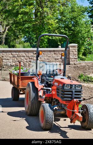 Ein roter Traktor mit einem Anhänger mit Blumen, der auf einer asphaltierten Straße in einem Gartenbereich mit grünen Bäumen und einer Steinmauer im Hintergrund geparkt ist. Stockfoto