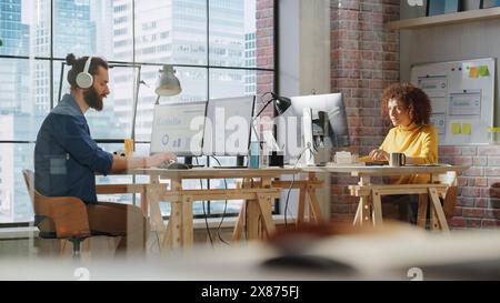 Mann und Frau sitzen hinter Schreibtischen neben dem großen Fenster mit Stadtblick und arbeiten mit analytischen Daten auf Computern. Zwei multikulturelle Kollegen arbeiten an der Erforschung und Entwicklung von Geschäftsstrategien. Stockfoto