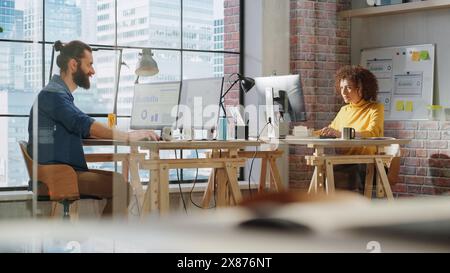 Zwei Manager sitzen hinter Schreibtischen neben dem großen Fenster mit Stadtansicht und arbeiten mit Analyseberichten auf einem Computer. Stilvolle männliche und multiethnische Frauen arbeiten an einem Projekt in einem Kreativbüro. Stockfoto