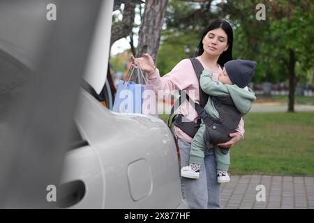 Die Mutter hält ihr Kind in der Tragetasche, während sie im Freien Einkaufstaschen in den Kofferraum legt Stockfoto