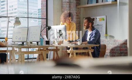 Zwei Manager sitzen hinter Schreibtischen neben dem großen Fenster mit Stadtansicht und arbeiten mit Berichten zu Marketingkampagnen auf einem Computer. Stilvolle männliche und multiethnische Frauen arbeiten an einem Projekt in einem Kreativbüro. Stockfoto