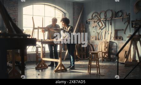 Erwachsene Zimmerfrau und eine junge multiethnische weibliche Auszubildende arbeiten im Loft Studio an einem neuen Stuhl Design. Inhaber kleiner Unternehmen sprechen über die Arbeit in einer stilvollen Möbelwerkstatt. Stockfoto