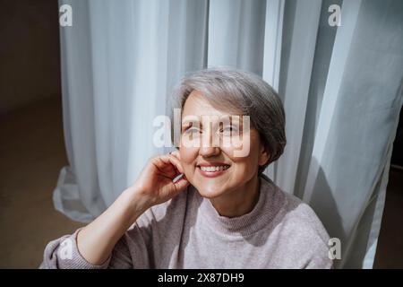 Nachdenklich lächelnde Frau vor dem Vorhang zu Hause Stockfoto