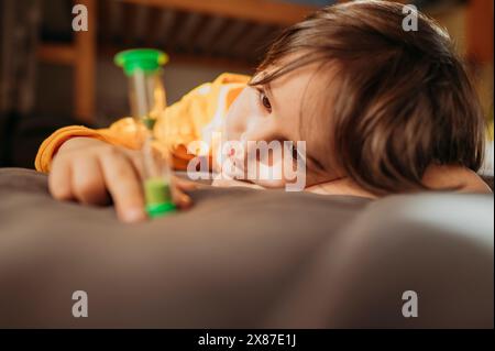 Nachdenklicher Junge, der auf Sanduhr im Bett blickt Stockfoto