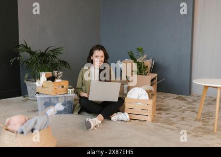 Lächelnde Frau mit Laptop, die in einem neuen Zuhause zwischen Boxen sitzt Stockfoto