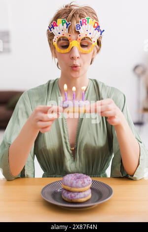 Frau in Geburtstagsgläsern, die zu Hause Kerze auf Donut bläst Stockfoto