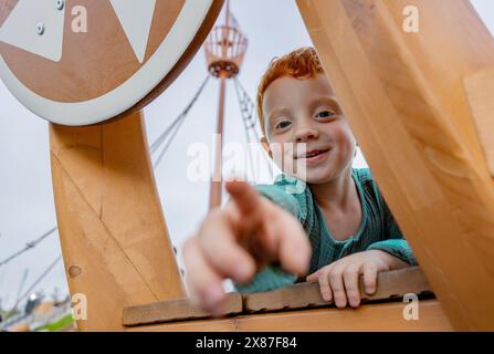 Ein lächelnder rothaariger Junge, der in die Nähe eines Holzobjekts auf den Spielplatz zeigt Stockfoto