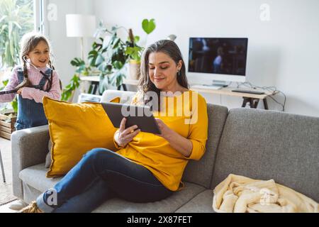 Lächelnde reife Frau, die einen Tablet-PC benutzt und zu Hause in der Nähe der Enkelin auf dem Sofa sitzt Stockfoto