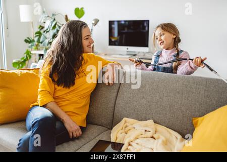 Lächelnde Frau, die auf dem Sofa sitzt und die Enkelin ansieht, die mit Stethoskop im Wohnzimmer spielt Stockfoto