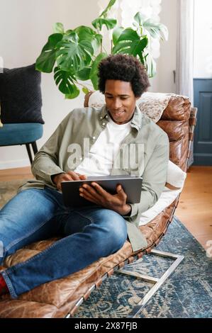 Lächelnder Geschäftsmann mit Laptop, der sich auf einem Klubsessel zurücklehnen und zu Hause arbeitet Stockfoto