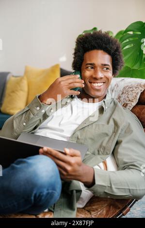 Glücklicher Geschäftsmann, der sich über das Handy zurücklehnt und mit Laptop auf dem Liegestuhl sitzt und zu Hause arbeitet Stockfoto