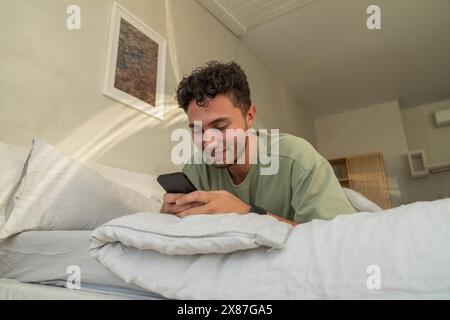 Lächelnder junger Mann, der zu Hause auf dem Bett liegt und das Smartphone benutzt Stockfoto