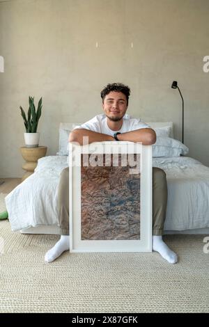 Ein lächelnder junger Mann sitzt zu Hause mit Bilderrahmen auf dem Bett Stockfoto