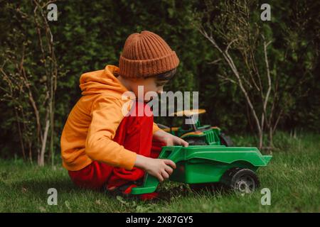 Junge, der mit einem grünen Spielzeugtraktor auf dem Gras im Hinterhof hockt und spielt Stockfoto