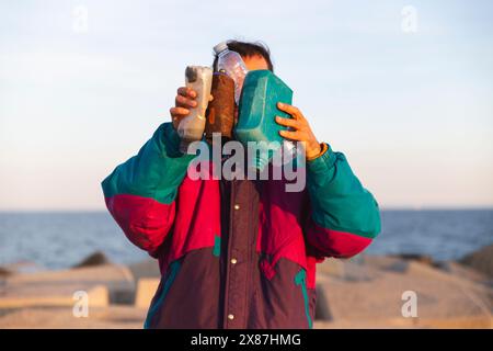 Mann in Jacke, der Plastikflaschen vor dem Gesicht hält, nahe dem Wellenbrecher Stockfoto