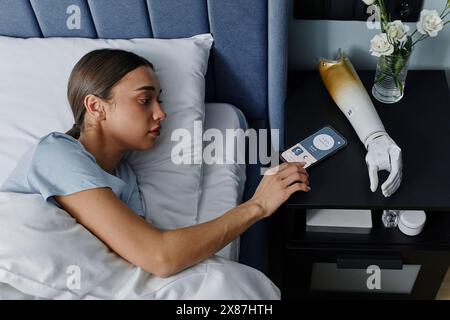 Junge Frau, die den Alarm auf dem Smartphone durch einen bionischen Arm im Schlafzimmer ausschaltet Stockfoto