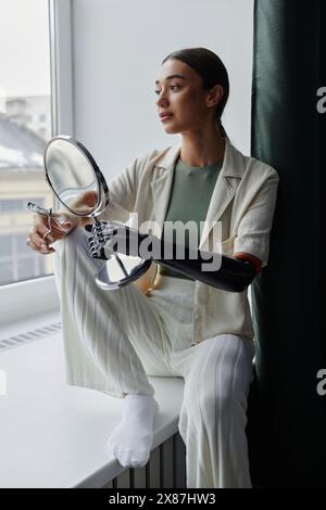 Nachdenkliche junge Frau mit bionischem Arm, der Spiegel hält und Wimpernlockenstab auf Fensterbank sitzt Stockfoto
