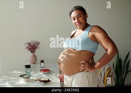 Lächelnde schwangere und große Frau, die zu Hause am Tisch steht Stockfoto