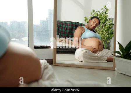 Eine schwangere Frau in Übergröße, die auf den Spiegel blickt und auf dem Boden im Zimmer sitzt Stockfoto