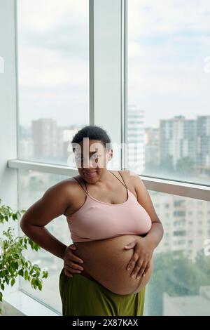 Lächelnde und große schwangere Frau, die zu Hause am Fenster steht Stockfoto