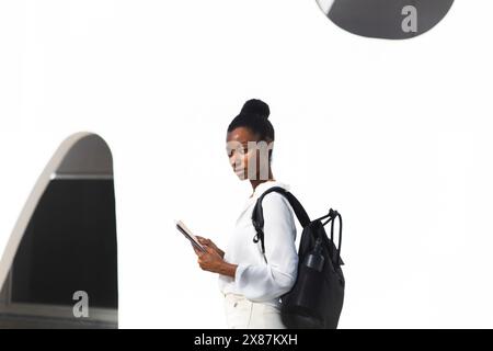 Selbstbewusste reife Frau mit Rucksack und Buch vor dem weißen Gebäude Stockfoto