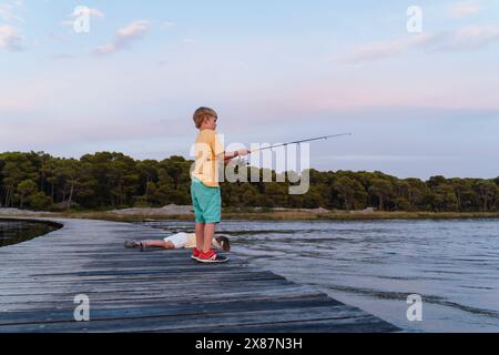 Bruder mit Angelrute bei Schwester, der bei Sonnenaufgang auf dem Pier über dem See liegt Stockfoto