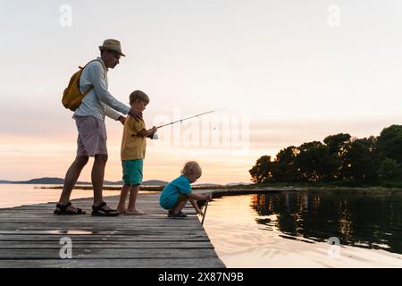 Vater mit Söhnen, die bei Sonnenaufgang am Pier über dem See angeln Stockfoto