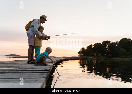 Vater mit Söhnen, die bei Sonnenaufgang am Pier über dem See angeln Stockfoto