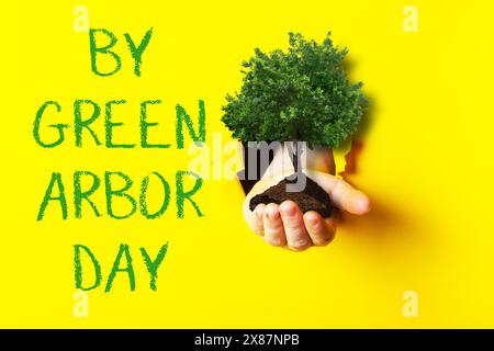 Eine Hand, die einen Baum vor einem gelben Hintergrund hält, mit den Worten von Green Arbor Day unten Stockfoto