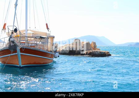 Die Yacht mit gefaltetem Segel mit weißer und brauner Schaluppe ist an klaren sonnigen Tagen auf dem Felsen im offenen Meer verankert. Deck auf Bug, Mast und Segeln in ruhiger See Stockfoto