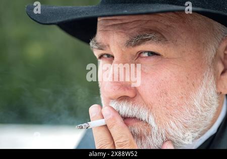 Nahaufnahme Porträt eines seriösen, stylischen Hipster-Großvaters im Ledermantel, der draußen raucht. Älterer Großvater, Großvater Rentner, Rentner-Konzept. Stockfoto