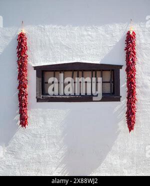 Seitliche Beleuchtung der roten Chili Ristras, die an der weißen Wand mit schwarzem Holzfenster an der Außenseite der traditionellen südNew Mexico Wohnstätte in Mesilla, NM, hängen Stockfoto