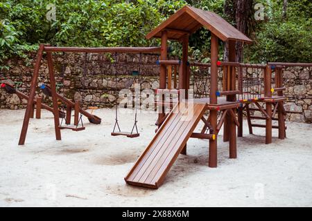 Ein gemütlicher Kinderspielplatz in einem Park, mit einer Holzrutsche, Schaukeln und einem Klettergerüst umgeben von üppigem Grün und einem Rusti Stockfoto