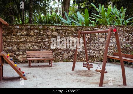 Ruhiger Kinderspielplatz in einem tropischen Park mit Holzbank, Schaukel und farbenfroher Kletterstruktur. Üppiges Laub und Histos Stockfoto