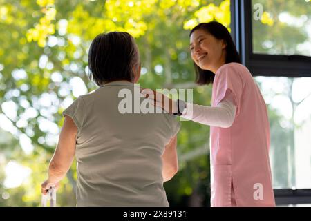 Zu Hause, asiatische junge Krankenschwester und Patientin mittleren Alters, in der Nähe des Fensters Stockfoto