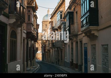 Eine enge Straße in Valletta, Malta, im Schatten, mit einer Kirchenkuppel im Sonnenlicht dahinter. Historische Architektur unter warmem Abendlicht Stockfoto