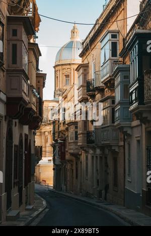 Eine enge Straße in Valletta, Malta, im Schatten, mit einer Kirchenkuppel im Sonnenlicht dahinter. Historische Architektur unter warmem Abendlicht Stockfoto