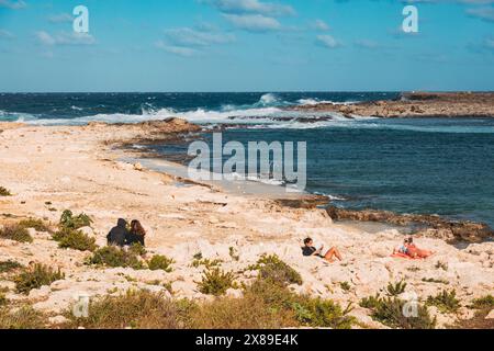 Sonnenanbeter entspannen und lesen in der Sonne an den felsigen Ufern von Qawra Point Beach, St. Paul's Bay, Malta Stockfoto