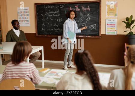 Moderner schwarzer Teenager, der an der Schule Englisch-Unterricht besucht, an der Tafel steht und Übungen macht, Kopierraum Stockfoto