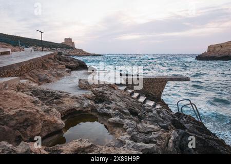 Wellen brechen gegen felsiges Gelände, während eine Steinplattform hinunter zum klaren Mittelmeer in Xlendi Beach, Munxar, Gozo, Malta führt Stockfoto