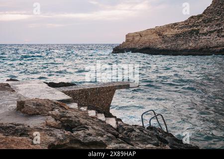 Wellen brechen gegen felsiges Gelände, während eine Steinplattform hinunter zum klaren Mittelmeer in Xlendi Beach, Munxar, Gozo, Malta führt Stockfoto