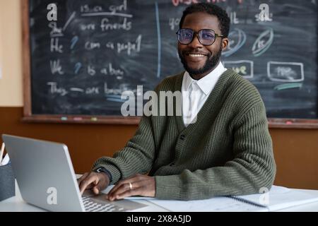 Mittleres Porträt eines jungen afroamerikanischen Englischlehrers mit Brille, der am Schreibtisch im Klassenzimmer sitzt und in die Kamera lächelt, während er einen Laptop benutzt Stockfoto