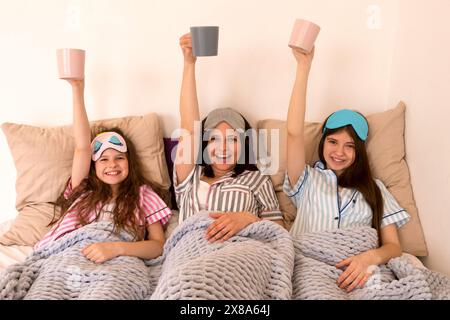Mutter und Töchter feiern mit erhabenen Tassen im Bett. Gemeinsame Familienzeit Stockfoto