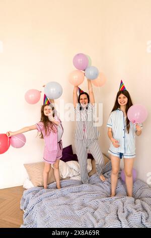 Familie in Partyhüten, die zu Hause mit Ballons auf dem Bett feiern. Schlafballon-Pyjama-Party Im Schlafzimmer Stockfoto