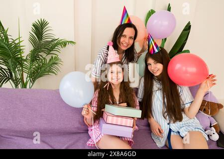 Mutter und Töchter feiern Geburtstag mit Ballons, Geschenken und Lächeln auf einer lila Couch. Stockfoto