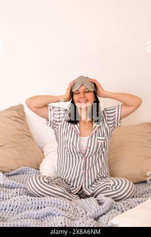 Frau im grauen gestreiften Pyjama, die Augenmaske anpasst, lächelt im Bett. Stockfoto